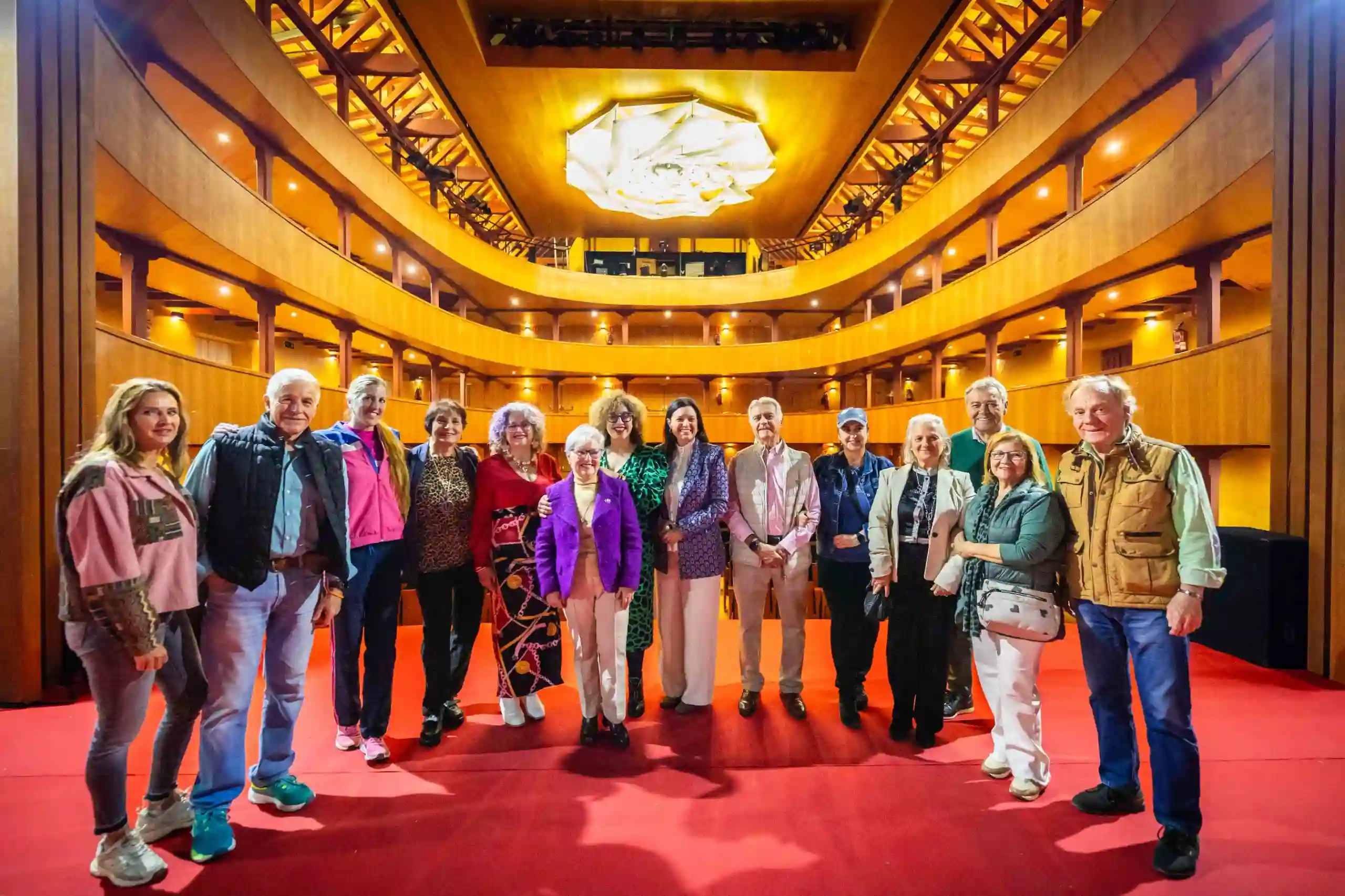 “Grupo de seis personas posando en un teatro iluminado; una mujer en el centro habla al micrófono durante un acto oficial.