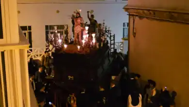 Procesión de Semana Santa en una calle estrecha con un paso iluminado por cirios y figuras religiosas rodeado de público.