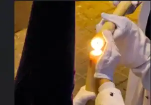 Manos con guantes blancos sosteniendo una gran vela doble encendida durante una procesión nocturna.