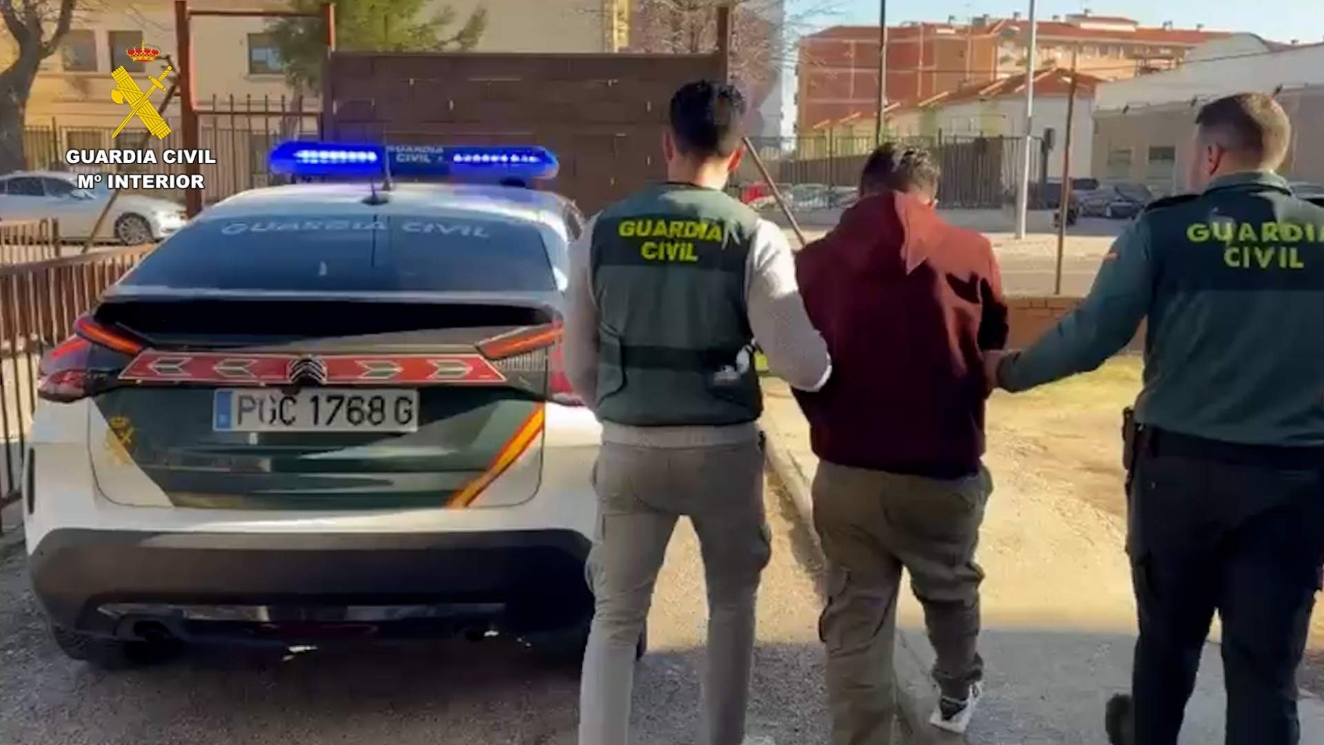 Dos agentes de la Guardia Civil conducen a un detenido, que lleva una sudadera granate, hacia un coche patrulla aparcado en la calle. El detenido va esposado y escoltado por un agente con chaleco reflectante y otro de uniforme. El texto es descriptivo y relevante para la noticia.