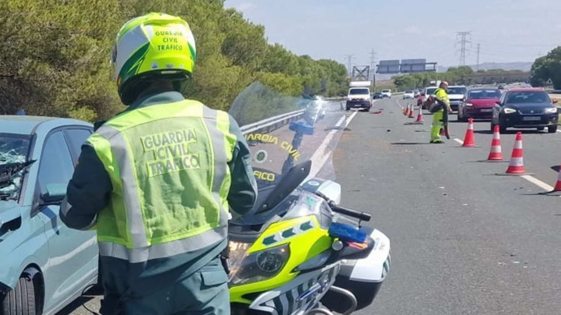 Un agente de la Guardia Civil de Tráfico de espaldas junto a una moto oficial regulando la circulación tras un accidente de tráfico.