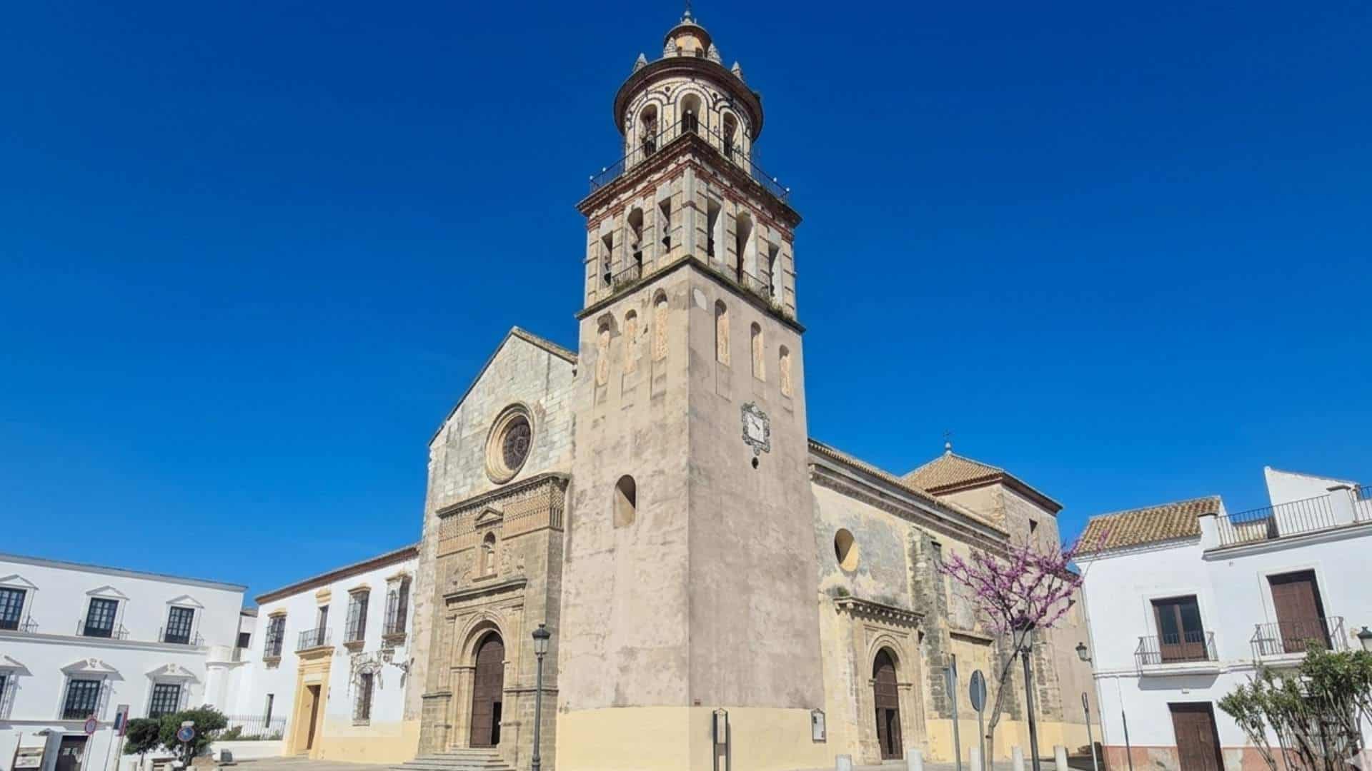 Fachada principal y torre campanario de la Parroquia de Nuestra Señora de la O en Sanlúcar de Barrameda bajo un cielo azul despejado.