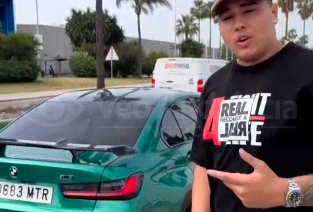 Joven frente a un coche BMW verde con matrícula española, señalando hacia el vehículo en una calle con palmeras.