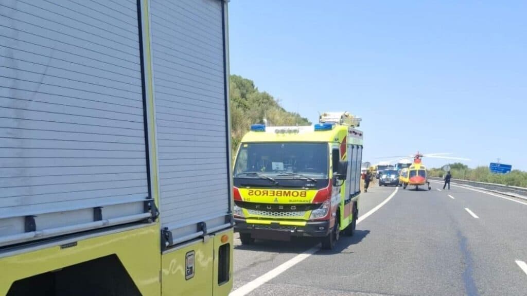 Vehículos de bomberos estacionados en una carretera con un helicóptero de emergencias aterrizado para atender un accidente.