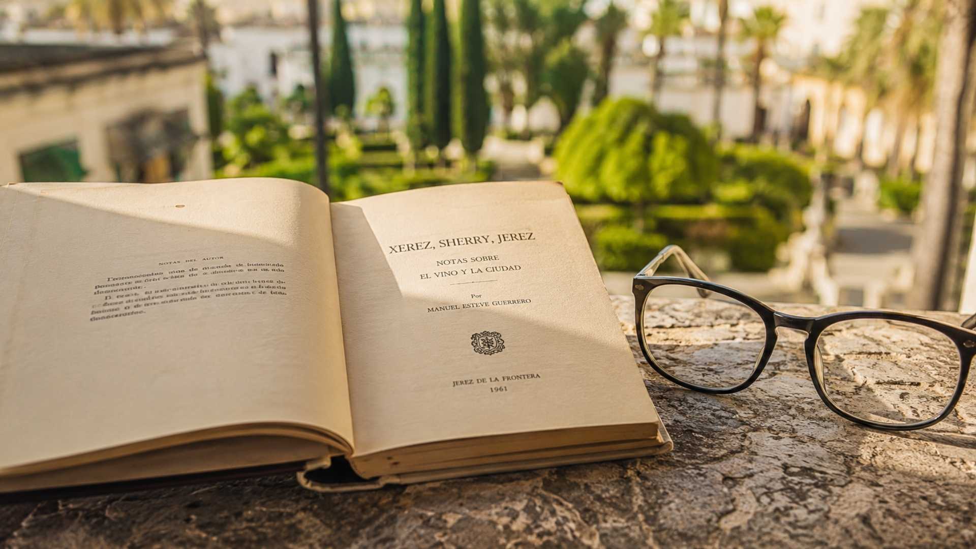 Libro antiguo y gafas sobre una mesa de piedra con jardín jerezano desenfocado al fondo.