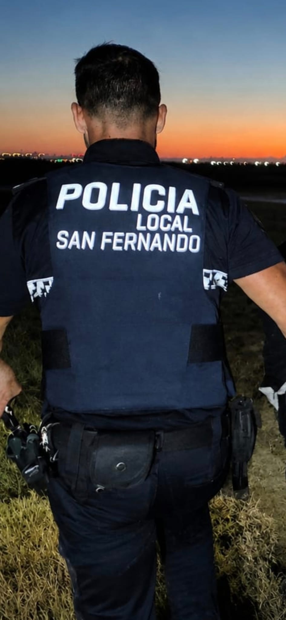Vista trasera de un agente de la Policía Local de San Fernando, Cádiz, uniformado con chaleco antibalas donde se lee 'POLICIA LOCAL SAN FERNANDO' durante un servicio al atardecer con cielo naranja.