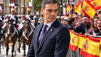 Primer plano de un micrófono de prensa frente a una multitud desenfocada durante un acto público, en referencia a los abucheos a Pedro Sánchez.