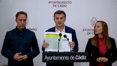 Tres personas durante una comparecencia en el Ayuntamiento de Cádiz; la persona situada en el centro sostiene un documento frente a varios micrófonos.