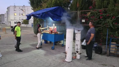 Hombre mostrando un recipiente metálico con castañas asadas junto a un puesto de venta bajo una estructura azul.