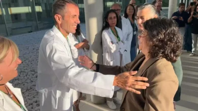 Varias personas conversan frente a un edificio moderno; un hombre con bata blanca saluda a una mujer con chaqueta marrón.