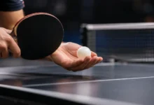 Primer plano de una pala de tenis de mesa y una pelota blanca sobre una mesa azul, con la red desenfocada al fondo.
