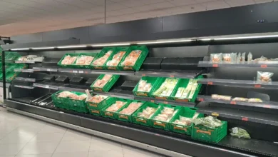 Estanterías refrigeradas casi vacías con algunos productos en los extremos y expositores de frutas y verduras al fondo de un supermercado.