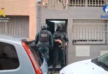 Dos agentes equipados con cascos y chalecos de la Guardia Civil junto a la entrada de un edificio, entre varios vehículos estacionados.
