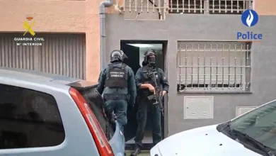 Dos agentes equipados con cascos y chalecos de la Guardia Civil junto a la entrada de un edificio, entre varios vehículos estacionados.