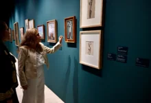 Dos mujeres observan obras de arte enmarcadas sobre una pared azul verdosa de una sala de exposiciones.