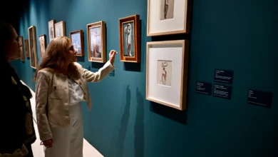 Dos mujeres observan obras de arte enmarcadas sobre una pared azul verdosa de una sala de exposiciones.
