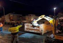Máquina excavadora amarilla junto a un contenedor metálico en una calle mojada durante la noche, con varias personas en la zona.