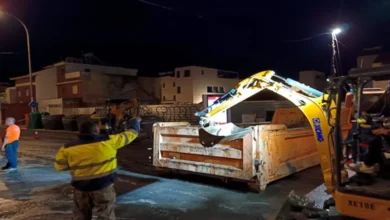 Máquina excavadora amarilla junto a un contenedor metálico en una calle mojada durante la noche, con varias personas en la zona.