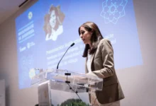 Mujer con traje beige interviene ante un micrófono desde un atril transparente, con una presentación proyectada al fondo.