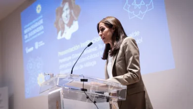 Mujer con traje beige interviene ante un micrófono desde un atril transparente, con una presentación proyectada al fondo.
