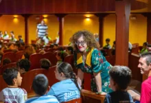 Una mujer con gafas y vestido verde de flores conversa con varios niños sentados en las butacas de un auditorio.