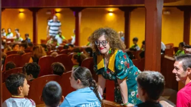 Una mujer con gafas y vestido verde de flores conversa con varios niños sentados en las butacas de un auditorio.