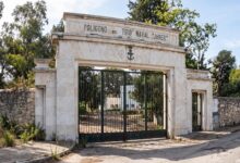 Entrada del Polígono de Tiro Naval Janer con portada de piedra blanca, verja metálica y árboles al fondo.