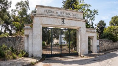 Entrada del Polígono de Tiro Naval Janer con portada de piedra blanca, verja metálica y árboles al fondo.