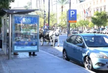 Mupi publicitario junto a una parada de autobús en una calle de Jerez, con un coche azul y un carruaje de caballos al fondo.