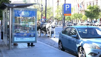 Mupi publicitario junto a una parada de autobús en una calle de Jerez, con un coche azul y un carruaje de caballos al fondo.