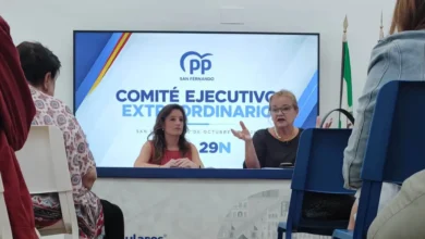 Dos mujeres sentadas detrás de una mesa durante una reunión, con varios asistentes en primer plano y una pantalla del PP de San Fernando al fondo.