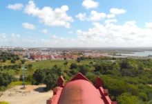 Vista panorámica de una zona arbolada con una cruz azul, una cubierta roja en primer plano, viviendas y agua al fondo.