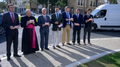 Siete hombres con vestimenta formal, entre ellos uno con indumentaria religiosa negra y fajín fucsia, permanecen de pie al aire libre sosteniendo papeles.