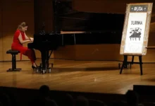 Una pianista vestida de rojo toca un piano de cola negro sobre un escenario de madera, junto a un cartel de la Ruta Turina.