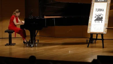 Una pianista vestida de rojo toca un piano de cola negro sobre un escenario de madera, junto a un cartel de la Ruta Turina.