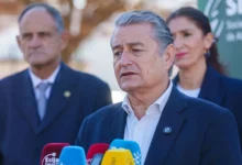 Un hombre de cabello gris y traje oscuro interviene ante varios micrófonos, con otras dos personas situadas detrás.
