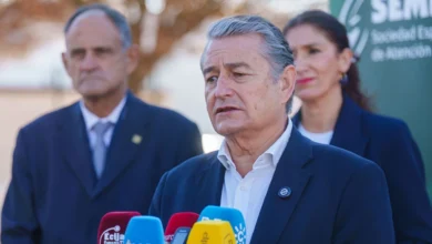 Un hombre de cabello gris y traje oscuro interviene ante varios micrófonos, con otras dos personas situadas detrás.