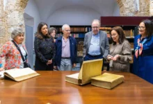 Varias personas observan libros antiguos abiertos y cerrados sobre una mesa de madera, delante de estanterías con volúmenes.