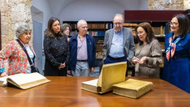 Varias personas observan libros antiguos abiertos y cerrados sobre una mesa de madera, delante de estanterías con volúmenes.