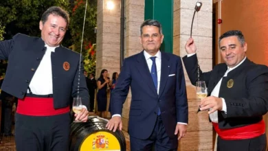 Tres hombres posan junto a una bota de vino con el rótulo «Embajada de España en Italia»; los dos situados a los lados visten chaquetas oscuras y fajines rojos, y sostienen copas y venencias.