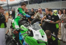 Piloto de motociclismo sobre una moto verde y blanca saluda a varias personas junto al paddock del circuito.