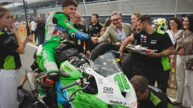 Piloto de motociclismo sobre una moto verde y blanca saluda a varias personas junto al paddock del circuito.