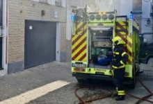 Un bombero junto a la parte trasera de un camión de extinción con mangueras desplegadas en una calle estrecha.