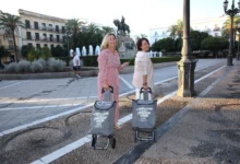 Dos mujeres posan junto a carros grises de la compra en una plaza, con una fuente y una escultura ecuestre al fondo.