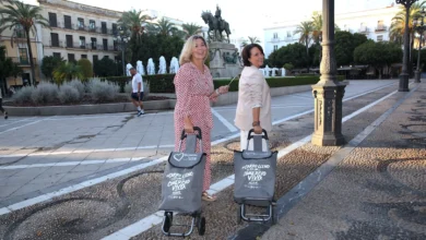 Dos mujeres posan junto a carros grises de la compra en una plaza, con una fuente y una escultura ecuestre al fondo.