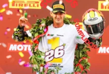 Piloto con gorra negra y camiseta blanca de campeón, decorada con un gran número uno, levanta un dedo y sostiene un casco durante una celebración con confeti.