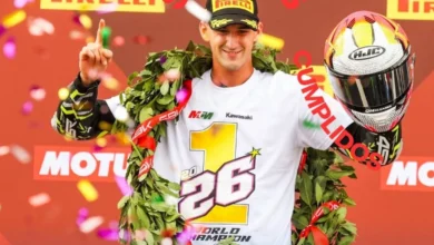 Piloto con gorra negra y camiseta blanca de campeón, decorada con un gran número uno, levanta un dedo y sostiene un casco durante una celebración con confeti.