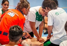 Varias jóvenes practican compresiones torácicas sobre un maniquí de reanimación mientras una profesional con ropa naranja del 061 supervisa la actividad.