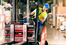 Trabajador con casco azul y chaleco amarillo sentado en una carretilla elevadora, rodeado de mercancías en un almacén.