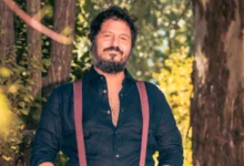 Hombre de cabello oscuro y barba, vestido con camisa negra y tirantes granates, posando al aire libre entre árboles.